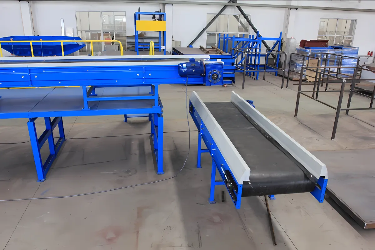 The image displays an industrial setting featuring a conveyor system as the central element. The conveyor system is designed to transport materials efficiently within the facility, likely as part of a larger manufacturing, processing, or sorting operation. Key Observations: Conveyor Belts: Two main conveyor belts are visible. The elevated belt with blue side rails is likely the primary conveyor, transporting materials over a longer distance or between different processing stations. The lower belt, perpendicular to the first, might be used for transferring materials to a different line or machine. Support Structure: The conveyor system is supported by a sturdy blue metal frame, providing stability and ensuring smooth operation. Motor and Drive System: An electric motor and drive components are visible, indicating the mechanism that powers the movement of the conveyor belts. Platform and Guardrails: A platform with yellow guardrails surrounds the starting point of the elevated conveyor, providing a safe working area for operators loading materials onto the belt. Industrial Environment: The open floor plan, high ceilings, and presence of other equipment in the background suggest a factory or industrial processing facility. Potential Applications and Functions: Material Handling: The conveyor system is likely used to transport various materials within the facility, such as raw materials, work-in-progress items, or finished products. Manufacturing and Assembly Lines: It could be part of a manufacturing or assembly line, where components are moved between different workstations for processing or assembly. Sorting and Distribution Centers: The system might be employed in sorting or distribution centers to move packages or items for further processing or shipping. Recycling Facilities: Conveyor systems are often used in recycling facilities to transport different types of materials for sorting and processing. Benefits of Conveyor Systems: Efficiency: Conveyor systems provide efficient and automated material handling, reducing manual labor and improving productivity. Continuous Flow: They allow for a continuous flow of materials, optimizing production or processing speeds. Versatility: Conveyor systems can be customized to handle various types of materials and adapted to different facility layouts. Safety: Automated conveyor systems can improve workplace safety by reducing manual handling and the risk of injuries. Overall, the image showcases a fundamental component of industrial material handling, emphasizing efficiency and automation within a production or processing environment.