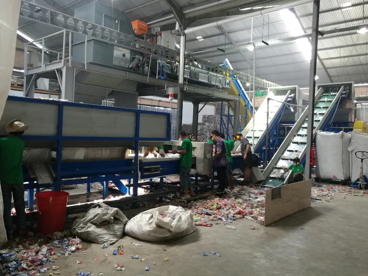 show the interior of a recycling facility, where workers are sorting through materials on a conveyor belt. This is likely a part of the initial sorting stage in a recycling process where workers separate different types of recyclable materials by hand. The facility seems to focus on the recycling of PET bottles, which are commonly used for beverages and other consumer products. The conveyor belt system is designed to move materials through the facility so that they can be sorted, cleaned, shredded, and eventually recycled into new products. The large bags and containers visible in the image suggest a collection of sorted materials ready for the next step in the recycling process. Manual sorting is a critical step in the recycling process as it ensures the purity of the materials being recycled, which is essential for high-quality recycling outcomes. The presence of workers in protective clothing, such as gloves and hats, indicates the emphasis on safety within the facility.