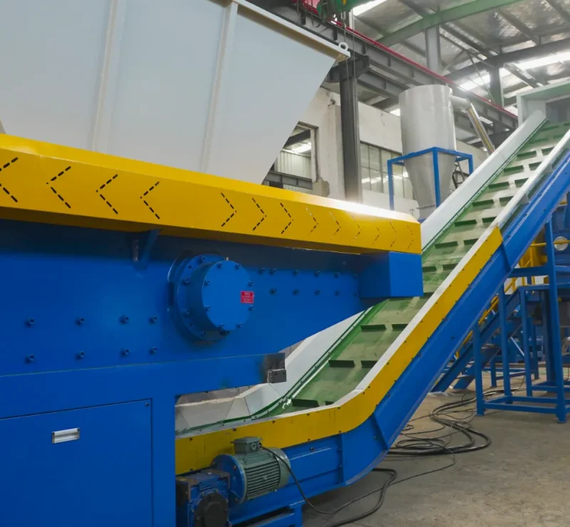 depicts a segment of industrial recycling equipment, possibly within a plastic recycling facility. The prominent blue machine with a yellow conveyor belt suggests it is part of a material handling system designed to transport plastic waste to different stages of the recycling process, such as sorting, washing, or shredding. The construction of the machinery indicates heavy-duty use, likely capable of handling large volumes of materials. The blue and yellow colors are not only aesthetic but also function as visual cues for safety and operational purposes. The staircase and platform structure in the background provide access to higher parts of the machinery for maintenance or to manage the recycling process. The presence of various hoppers and conduits suggests a sophisticated system to manage and direct the flow of materials through the recycling process. The investment in such machinery typically reflects a commitment to efficient, high-volume processing of recyclable materials to turn waste into reusable raw materials.