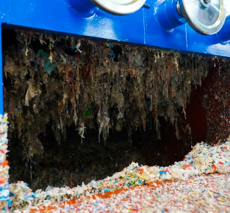 a close-up of a machine used in the recycling process, specifically what appears to be the interior of a shredder or grinder. Inside, we can see shredded plastic fragments, which are the result of the machine processing larger pieces of plastic waste into smaller, more manageable sizes. The shredding process is essential in the recycling of plastics as it prepares the material for further steps such as washing, separating, and eventually melting down to form new plastic products. The shredded plastic shown is varied in color, indicating that the machine has processed a mix of different types of plastic materials. The cleanliness of the blades and the interior of the machine is crucial for efficient operation and preventing contamination between different types of plastics, which can be critical for the quality of the recycled material. The machine appears to be in operation, with the shredded plastic exiting the machine, ready to move on to the next stage of recycling.