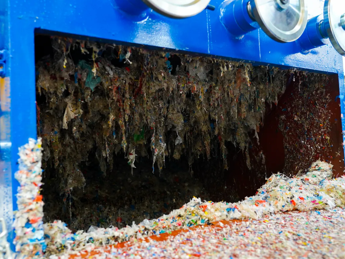 a close-up of a machine used in the recycling process, specifically what appears to be the interior of a shredder or grinder. Inside, we can see shredded plastic fragments, which are the result of the machine processing larger pieces of plastic waste into smaller, more manageable sizes. The shredding process is essential in the recycling of plastics as it prepares the material for further steps such as washing, separating, and eventually melting down to form new plastic products. The shredded plastic shown is varied in color, indicating that the machine has processed a mix of different types of plastic materials. The cleanliness of the blades and the interior of the machine is crucial for efficient operation and preventing contamination between different types of plastics, which can be critical for the quality of the recycled material. The machine appears to be in operation, with the shredded plastic exiting the machine, ready to move on to the next stage of recycling.