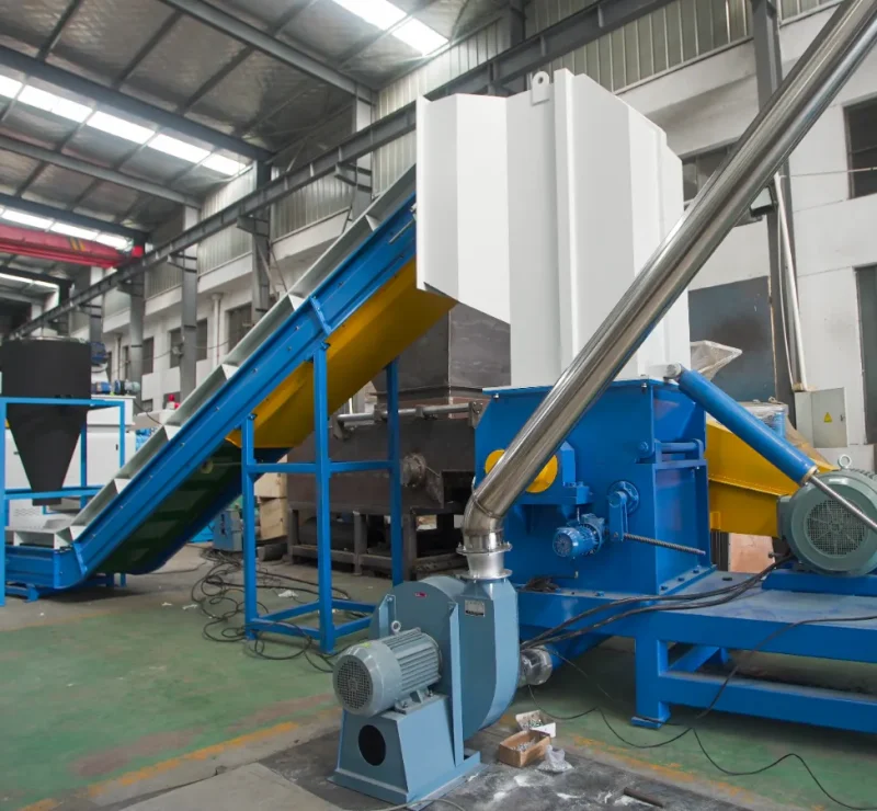 a plastic recycling machine within an industrial setting. The machine appears to include a conveyor belt system for transporting materials, which may be for feeding plastic waste into the machine. There's also a large hopper where materials can be stored before processing, and a series of pipes that likely transport materials or possibly air to separate waste products. The blue machinery suggests that this is part of a system designed for the shredding, washing, or pelletizing of plastics as part of the recycling process. Such systems are crucial in breaking down plastic waste into smaller, manageable pieces that can then be thoroughly cleaned, sorted, and eventually melted down and reformed into new plastic products. The electrical motors and metal framework indicate a robust construction designed to withstand the heavy, continuous loads typical of such industrial processes. Safety appears to be a consideration as well, with the motors and moving parts being well-guarded.