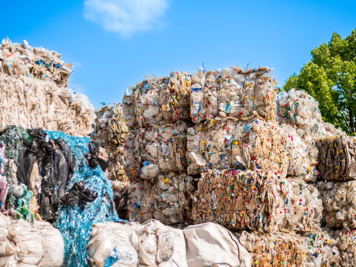 grandes fardos de residuos plásticos comprimidos, que normalmente se preparan para el reciclaje. Estas balas se forman después de los procesos de clasificación y limpieza, en los que diferentes tipos de plásticos se comprimen en bloques manejables para facilitar su transporte y procesamiento posterior. La variedad de colores dentro de cada fardo indica una mezcla de diferentes artículos de plástico, lo que sugiere que estos fardos pueden estar destinados a una instalación que clasificará y reciclará el material en nuevos productos plásticos. Esta es una práctica común en la industria del reciclaje para reducir el volumen de residuos, haciendo más rentable el transporte de materiales a las plantas de reciclaje. Las instalaciones de reciclaje suelen utilizar empacadoras para compactar los materiales reciclables clasificados en fardos densos y atados. Las pacas ahorran espacio durante el almacenamiento y el transporte, y su forma uniforme las hace más fáciles de manipular y procesar.