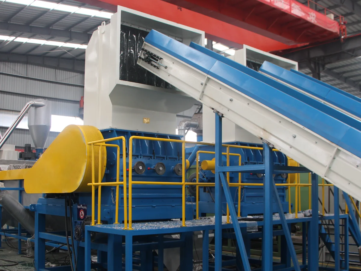 We can see a large blue machinery with a conveyor belt that seems to be leading to a grinding or washing unit. The yellow components might be safety guards, which are common in industrial settings to ensure worker safety. The machine underneath the conveyor could be a wet grinder, which is used to break down plastic waste into smaller pieces while simultaneously washing it. This process is a crucial step in recycling as it prepares the plastic for further purification and processing. The presence of water pipes and the design of the machinery suggest that the system is set up for wet grinding, a process which helps in removing contaminants and labels from the plastic waste before it moves to the next phase, which might include drying, additional sorting, or melting down into pellets for reuse.