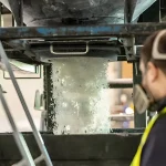 a worker observing an industrial process where a stream of plastic flakes or pellets is falling into what seems to be a collection bin or hopper. The worker is wearing personal protective equipment, including a high-visibility vest and hearing protection, which suggests a focus on safety in a potentially noisy environment. This scene could be part of a plastic recycling operation where shredded plastic materials are being processed further. After being washed and shredded into flakes, plastics are often sorted by type and color, then melted down and extruded into pellets, which serve as the raw material for manufacturing new plastic products. The machine from which the plastic materials are falling could be part of a pelletizing line, or it might be a part of a sorting system where materials are sifted and separated. The continuous flow of material indicates an automated and efficient recycling process, vital for handling large quantities of material in a recycling facility.