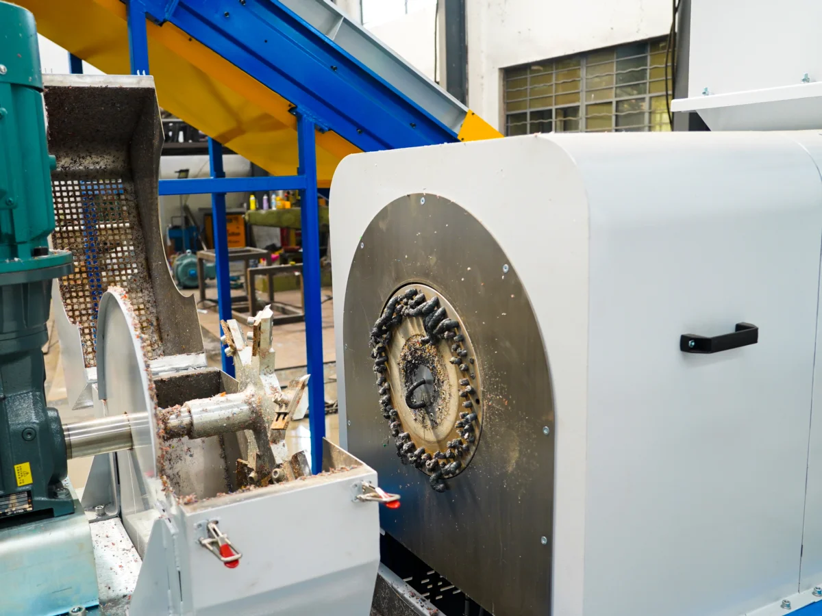 a part of a recycling line. Visible is a circular saw or cutting wheel covered with plastic debris, suggesting that it has been used to chop or grind plastic material. Such machinery is typically used to reduce the size of plastic waste to make it easier to process in later stages of recycling, like washing, pelletizing, or densification. The background includes more industrial equipment and a conveyor system, indicating this is part of a larger processing facility. Safety measures like guards and emergency stops seem to be in place, which are essential for operating heavy machinery.