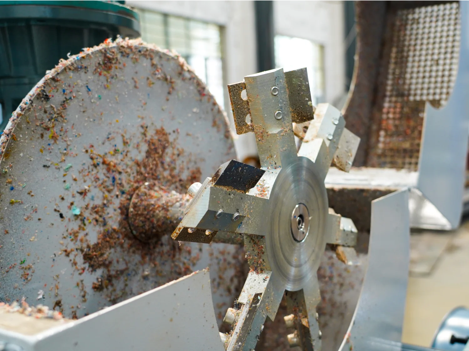The image appears to show part of a machine used for processing materials. It specifically features a metallic rotor or cutting blade covered in processed material, possibly indicating that it's part of a plastic granulator or shredding machine. This machinery is typically used in recycling or manufacturing settings to break down plastic or other materials into smaller granules for reuse or further processing. The worn and varied appearance of the material on the rotor suggests a collection of different shredded substances.