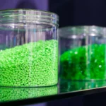 The picture shows a close-up of three glass jars containing colored plastic granules. The jar in the foreground is filled with bright green granules, while the jars in the background contain different shades of green and blue granules. These granules are typically used as raw material in the plastics industry for the manufacturing of various plastic products through processes like injection molding and extrusion.
