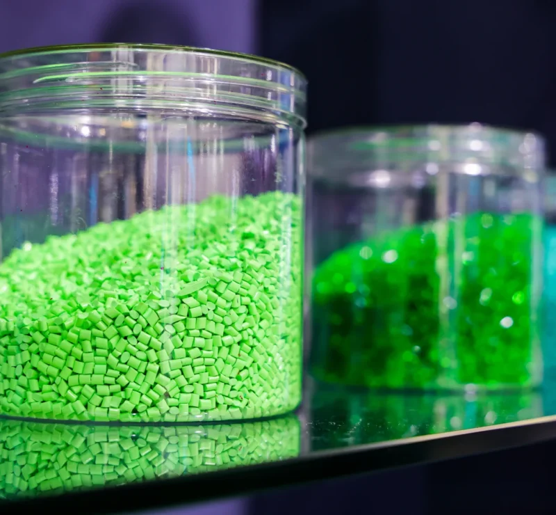 Slika prikazuje izbliza tri staklene posude koje sadrže plastične granule u boji. Staklenka u prvom planu ispunjena je svijetlo zelenim granulama, dok staklenke u pozadini sadrže različite nijanse zelenih i plavih granula. Ove se granule obično koriste kao sirovina u industriji plastike za proizvodnju raznih plastičnih proizvoda kroz postupke poput injekcijskog prešanja i ekstruzije.