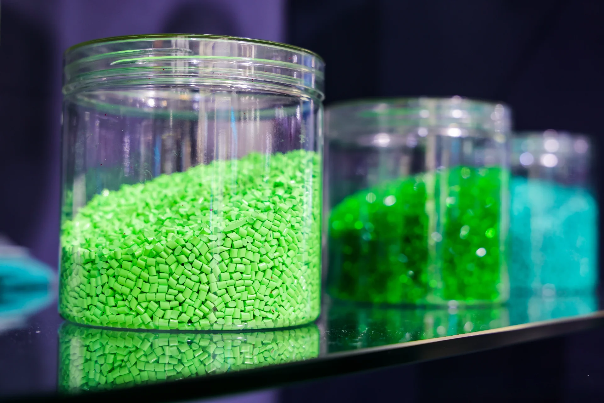 The picture shows a close-up of three glass jars containing colored plastic granules. The jar in the foreground is filled with bright green granules, while the jars in the background contain different shades of green and blue granules. These granules are typically used as raw material in the plastics industry for the manufacturing of various plastic products through processes like injection molding and extrusion.