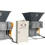 Dieses Bild zeigt zwei feststehende Einwellenzerkleinerer mit Schaufel. Jede Zerkleinerungseinheit verfügt über einen großen Trichter für die Materialzufuhr, einen Zerkleinerungsmechanismus, der an der Basis sichtbar ist, und ein angeschlossenes elektrisches Bedienfeld mit verschiedenen Tasten und Anzeigen. Die Geräte sind für den industriellen Einsatz konzipiert und können Materialien in kleinere Stücke für die Verarbeitung oder das Recycling zerkleinern. Die Maschinen sind robust und in den Farben Grau und Gelb gehalten, was die industrielle Stärke und den Zweck der Zerkleinerer unterstreicht.